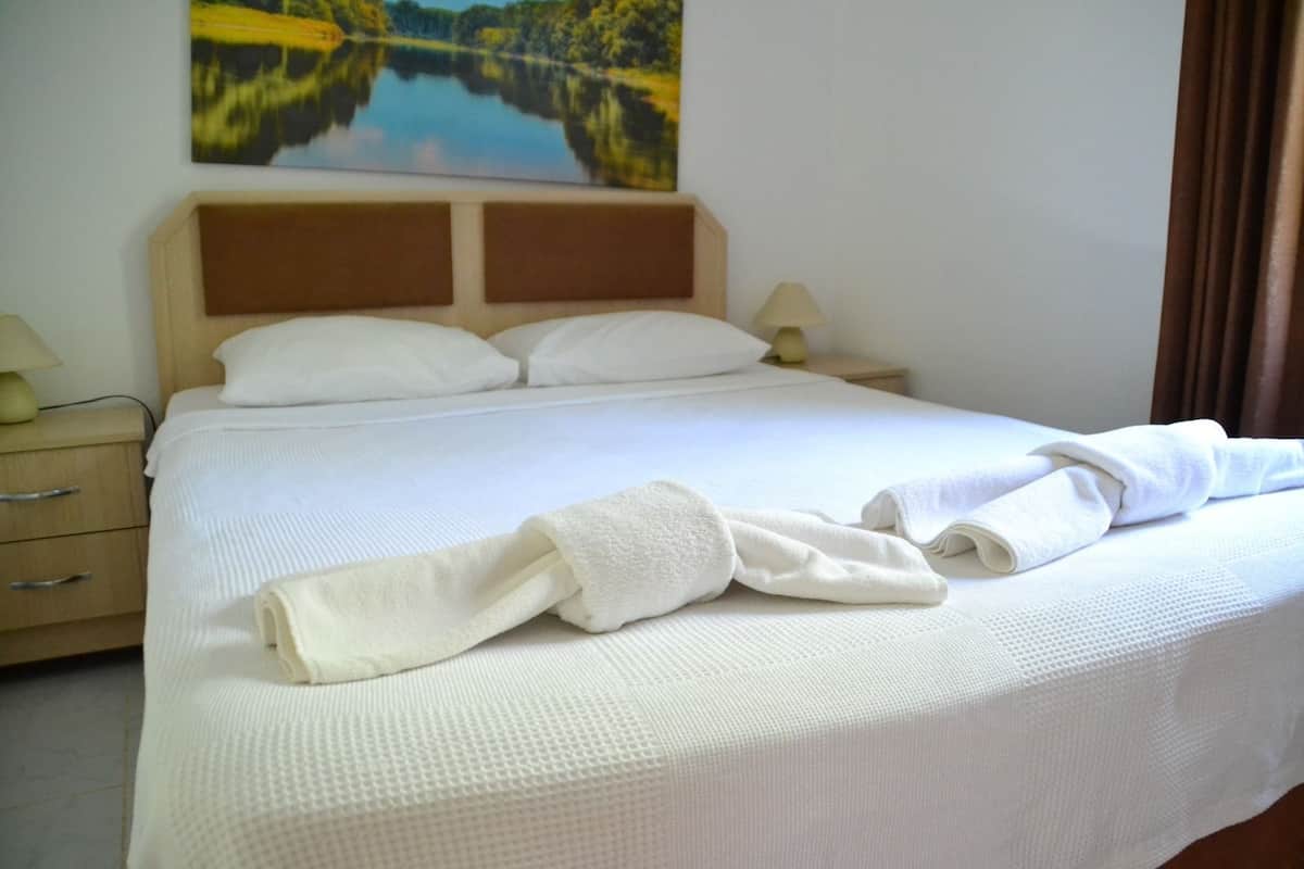 A neatly made bed with white linens, a wooden headboard, and a painting of a lake on the wall.