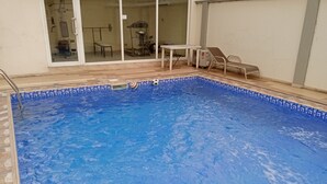 En swimmingpool med flisekant, en glasdør der fører til et rum med bord og stole, og en stol udenfor døren.