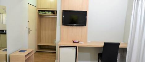 Quarto compacto com escrivaninha de madeira, mini-geladeira, TV fixada na parede e cadeira preta.