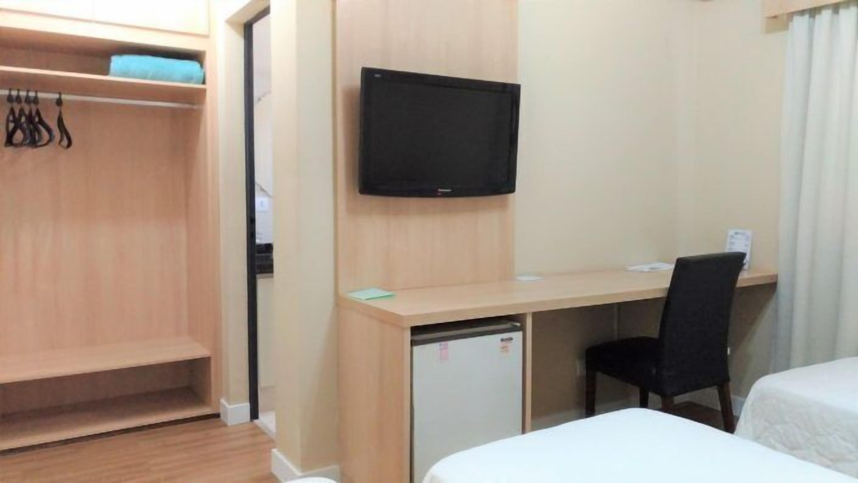 Quarto de hotel com escrivaninha de madeira, televisão montada acima, pequeno frigorífico, uma cadeira preta e duas camas.