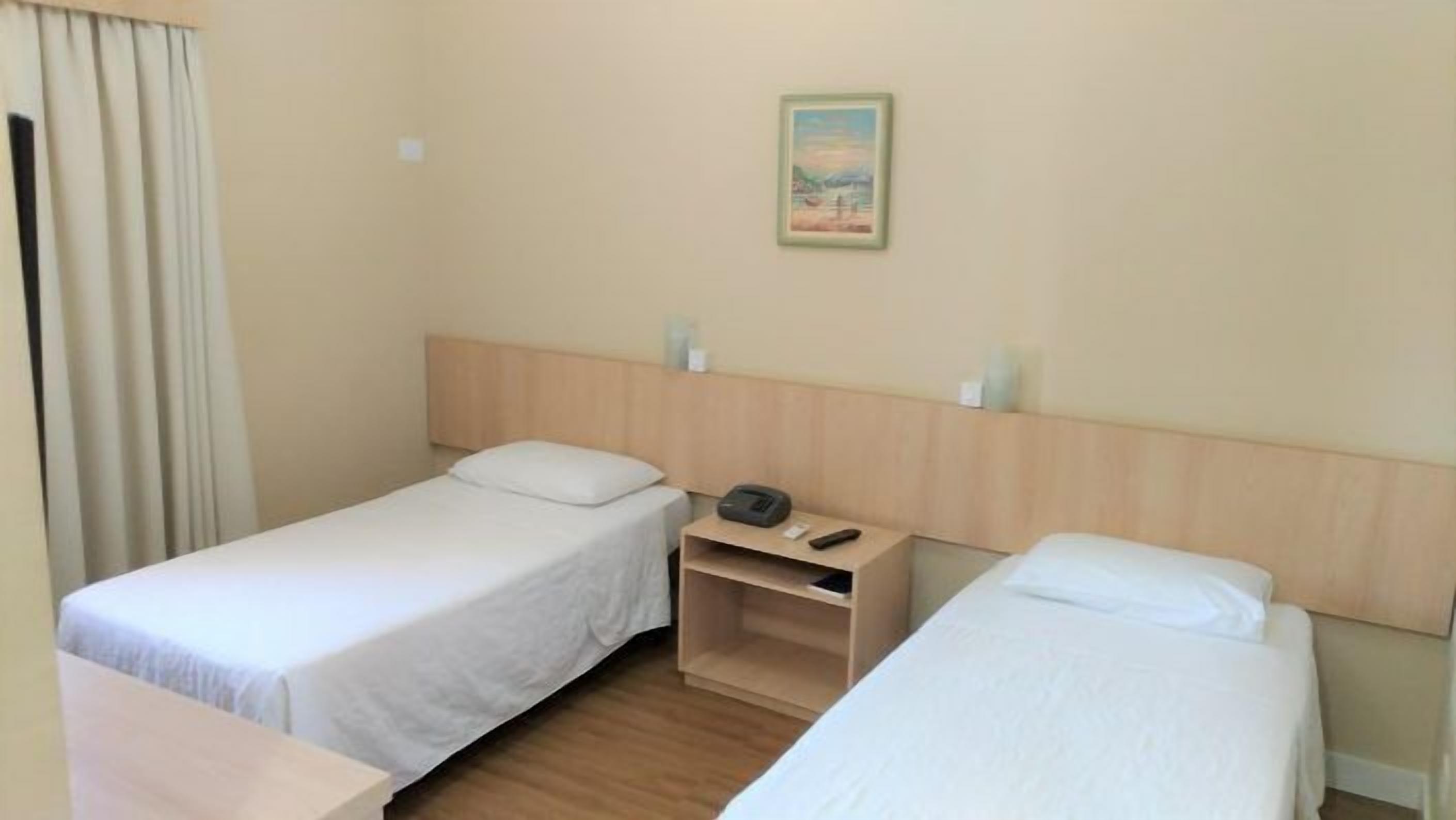 A hotel room with two single beds, a small wooden nightstand, a painting on the wall, and a window with curtains.