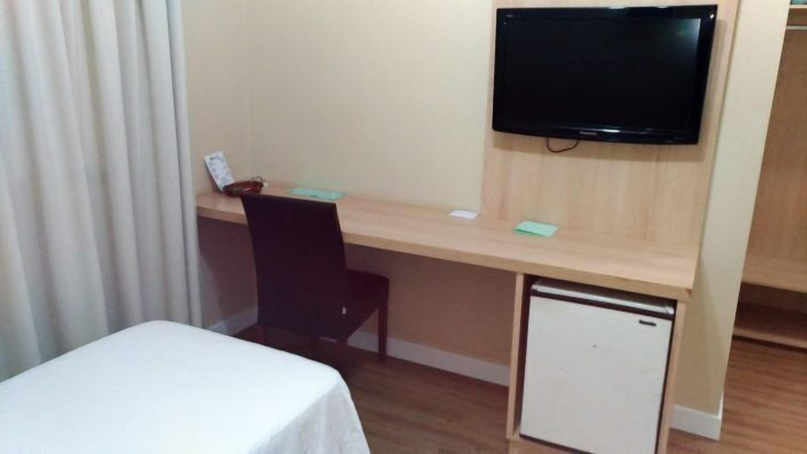 A compact hotel room with a single bed, a desk with a chair, a small refrigerator, and a flat-screen TV mounted on the wall.