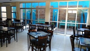 Een restaurant met meerdere tafels en stoelen, grote ramen met uitzicht op palmbomen en een betegelde vloer.