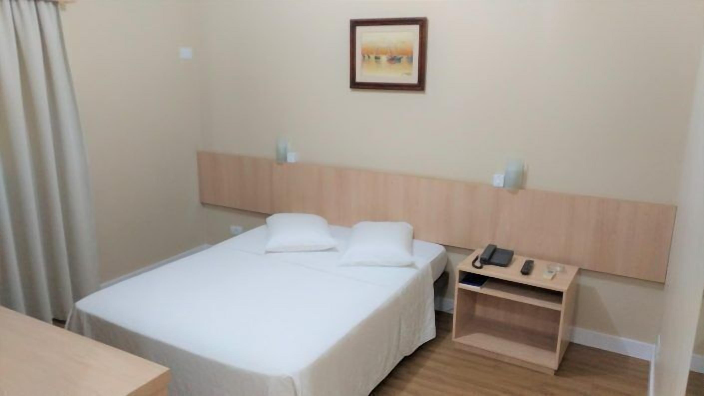 Una habitación de hotel con una cama, una mesita de noche con teléfono y control remoto, y un cuadro en la pared.