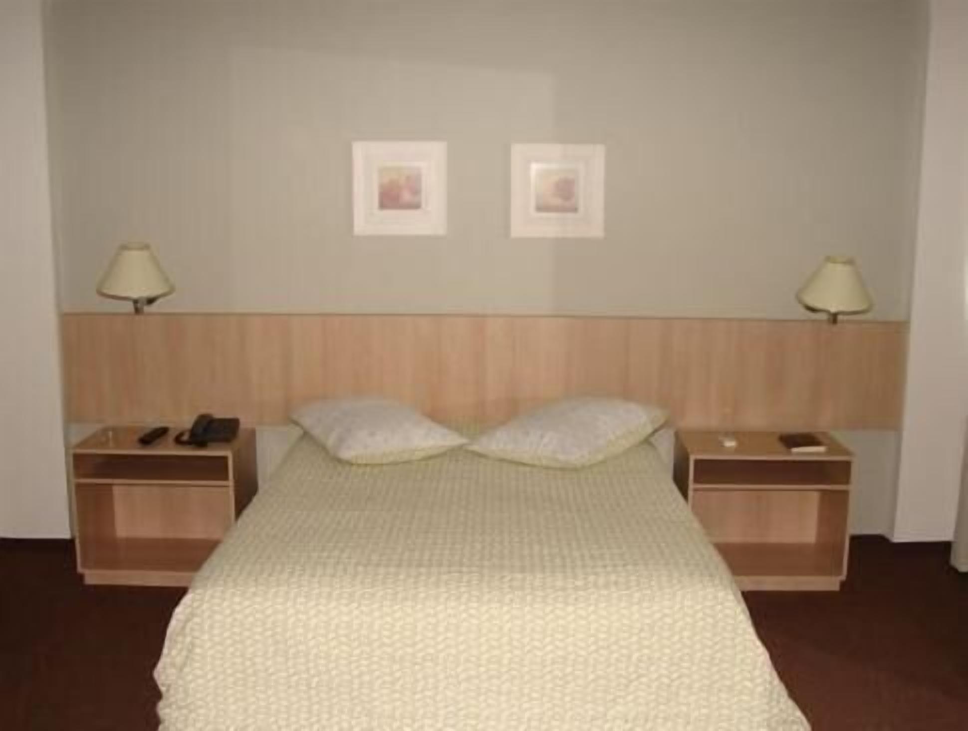 Quarto de hotel simples com uma cama, duas mesas de cabeceira, um abajur em cada lado e duas imagens emolduradas na parede.