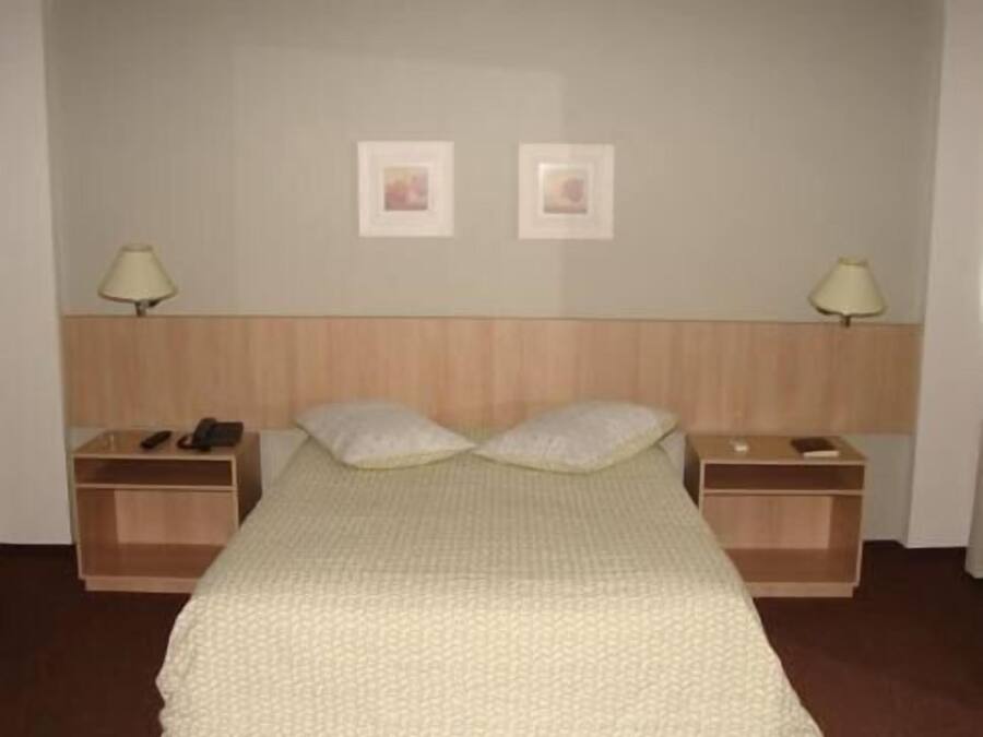 Quarto de hotel simples com uma cama, duas mesas de cabeceira, um abajur em cada lado e duas imagens emolduradas na parede.