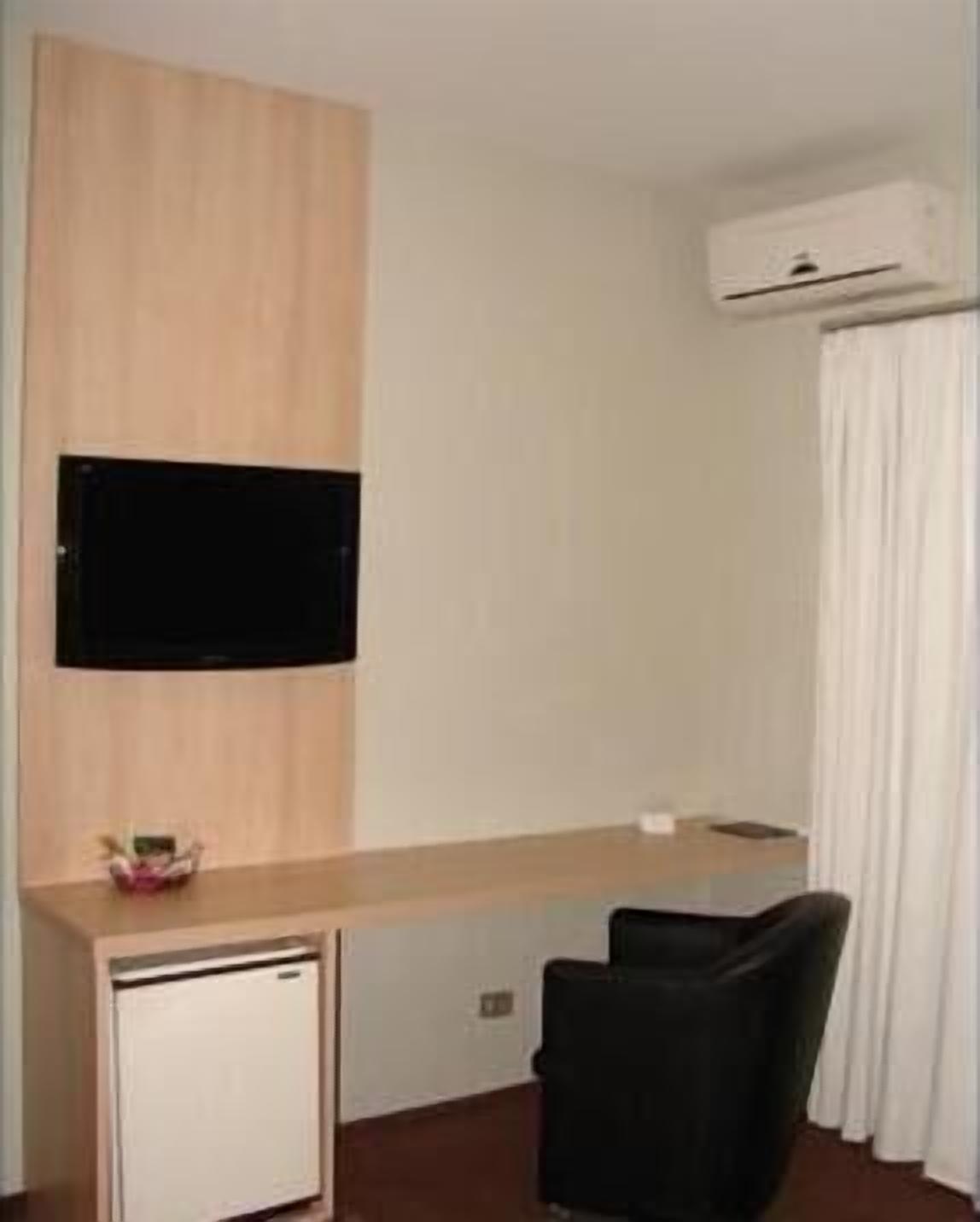 Una camera d'albergo con una scrivania in legno, un piccolo frigorifero bianco, una sedia nera e una TV a schermo piatto montata al muro.