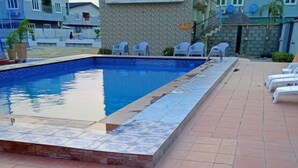 A rectangular swimming pool with a tiled edge, surrounded by a tiled deck. There are white plastic chairs arranged along the pool's edge. - The Gold (Lekki)