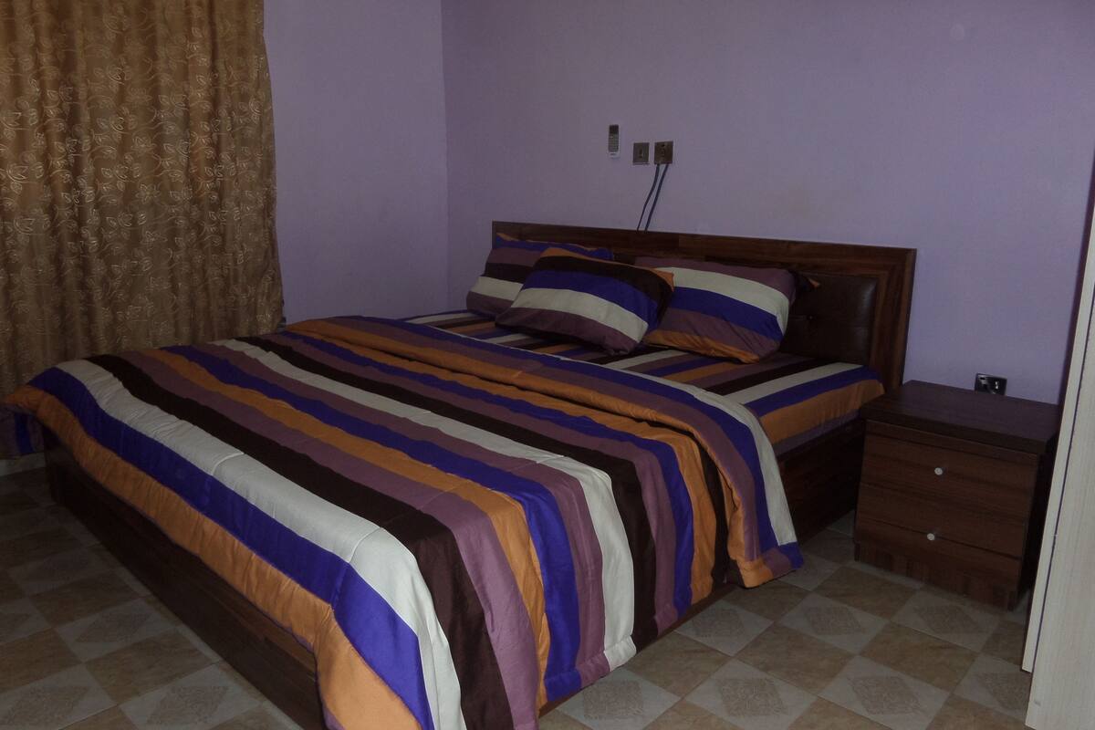 A bedroom with a bed, a nightstand, a drawer, a curtain, and a wall socket.