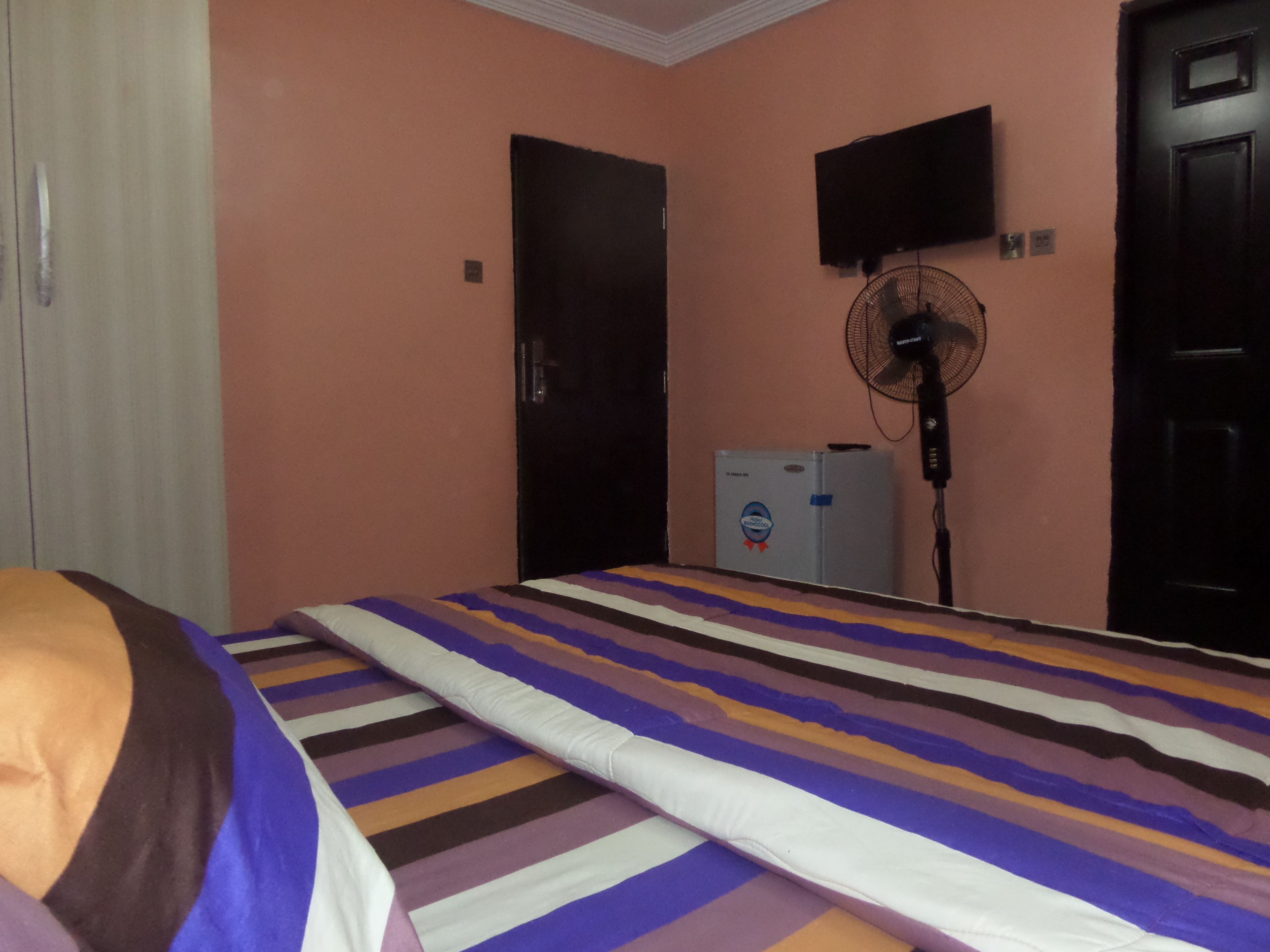 Un dormitorio con cama, ventilador, televisión y refrigerador.