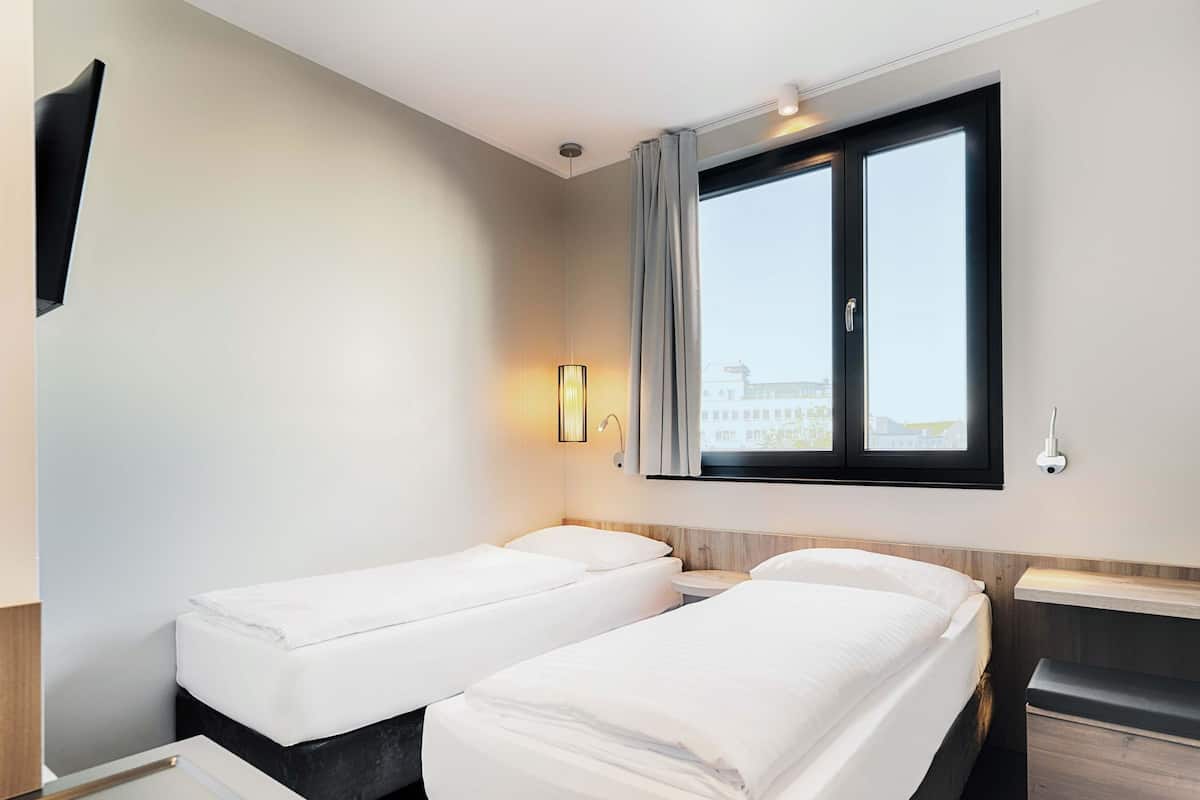 Une chambre d’hôtel avec deux lits simples, une fenêtre avec des rideaux, un téléviseur fixé au mur et une table de chevet avec une lampe.