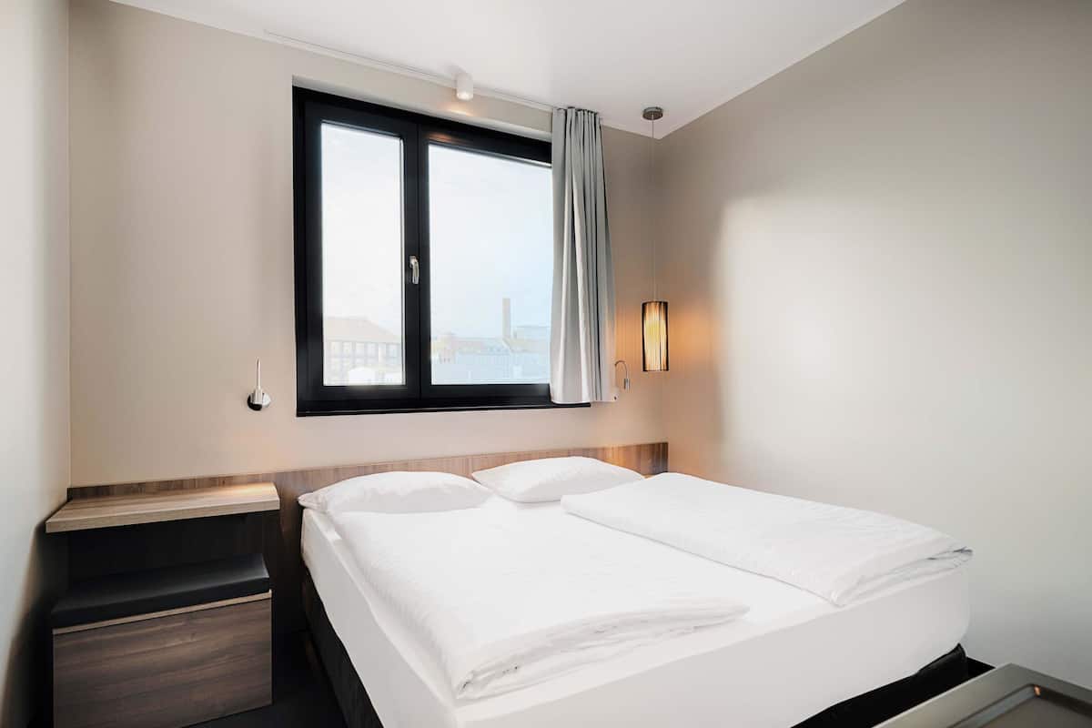 Une chambre d’hôtel avec un lit, une table de chevet, une fenêtre avec des rideaux et un luminaire fixé au mur.