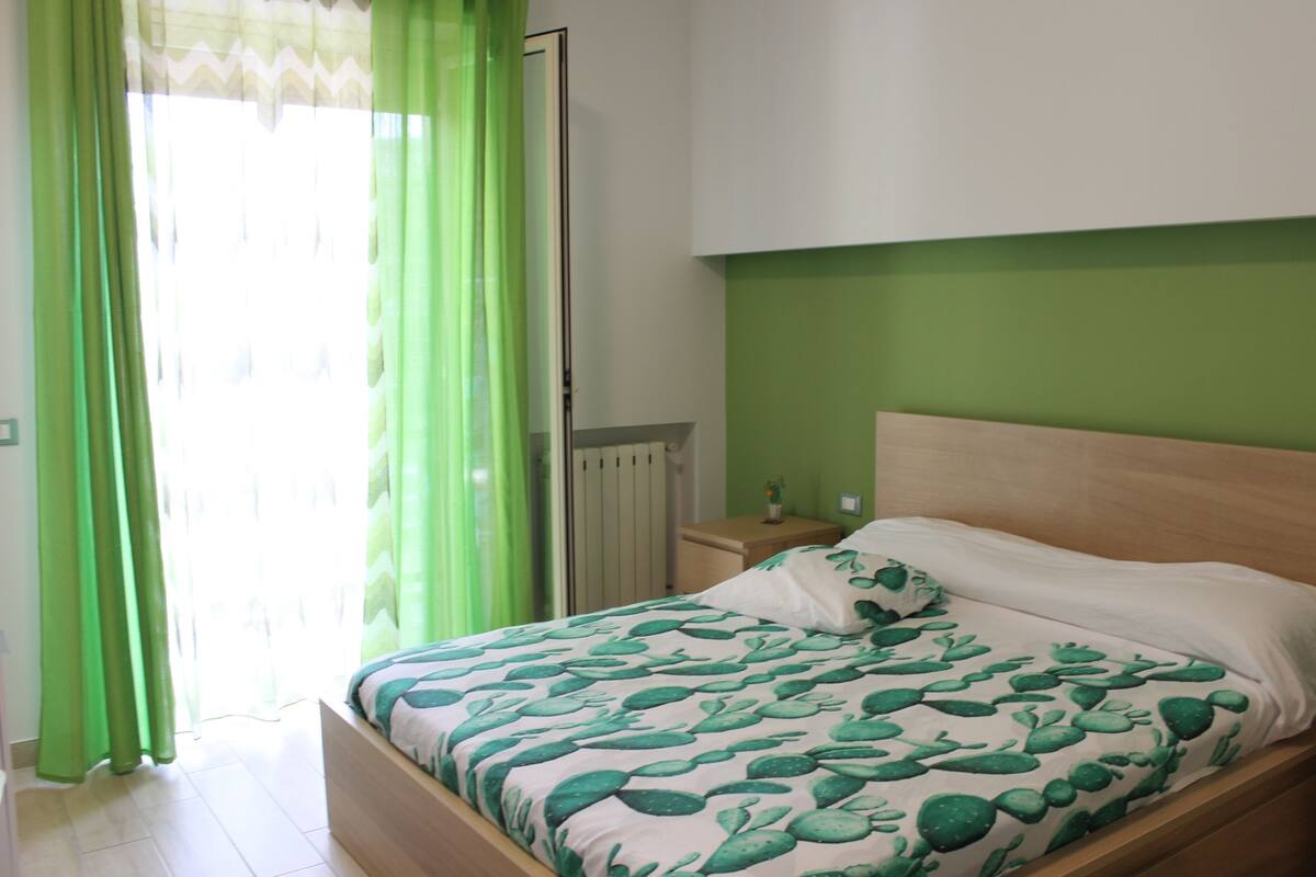 Un dormitorio con cama, cortinas verdes, un radiador y una mesita de noche con lámpara.
