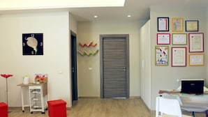 A modern room with a wooden floor, a desk with a laptop, a white chair, a red stool, a wall clock, and framed certificates.
