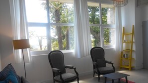 A living room with two patterned armchairs, a small table, a colorful rug, a yellow bookshelf, and large windows with curtains.