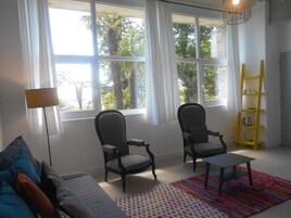 Una sala de estar con dos sillones estampados, una mesita, un tapete colorido, una estantería amarilla y ventanas grandes con cortinas.