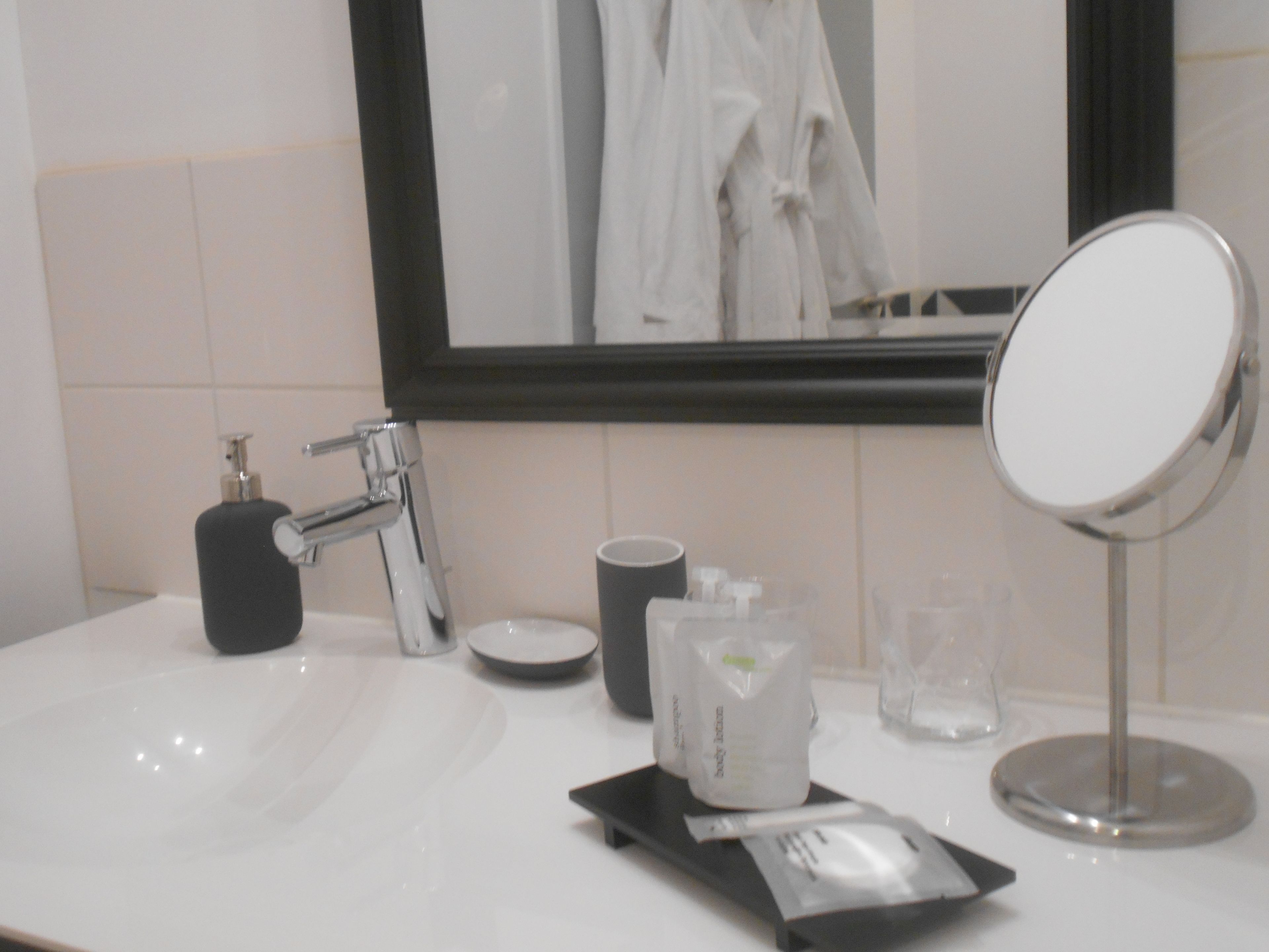 Un baño con lavamanos blanco, espejo, dispensador de jabón, taza y una pequeña bandeja con un paquete.