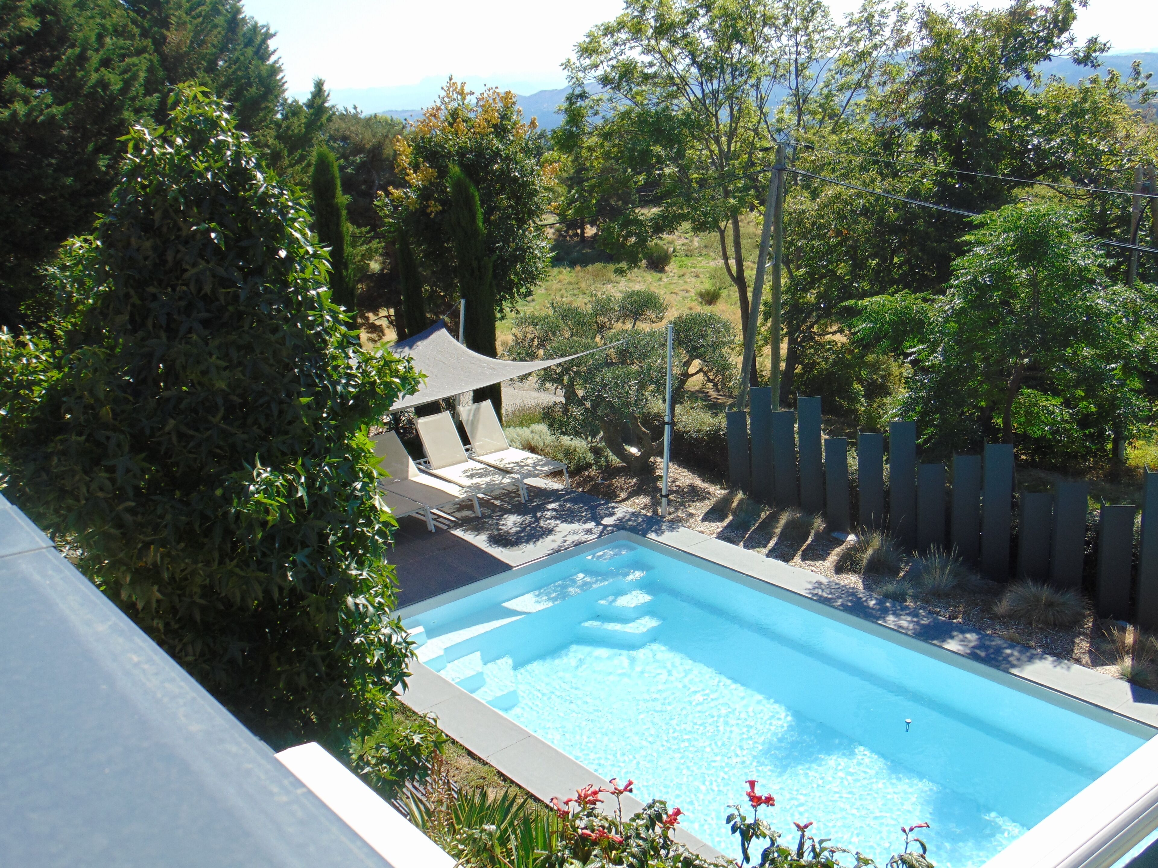 A swimming pool with a shaded lounge area, surrounded by a fence and greenery.