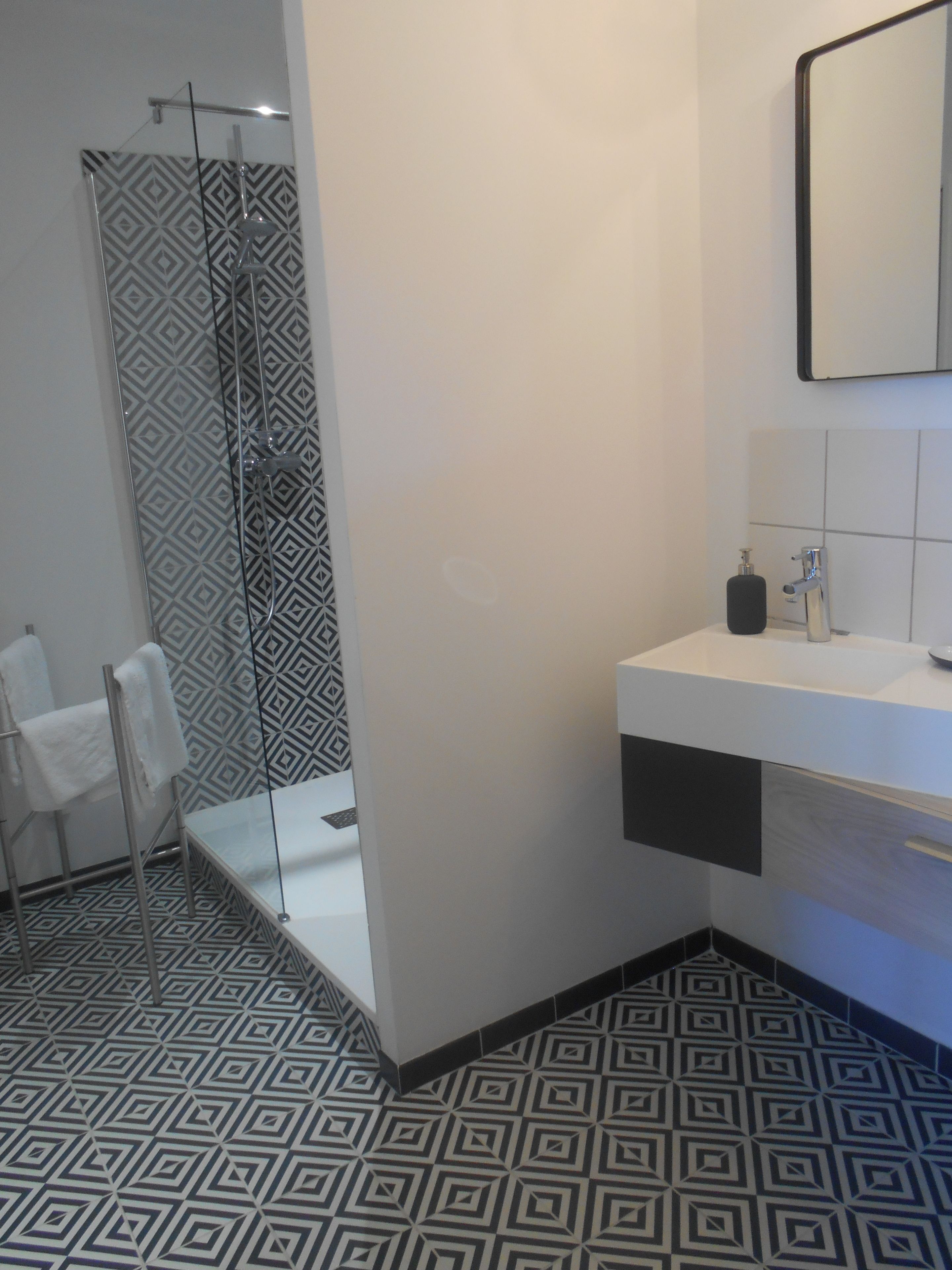 Un baño moderno con piso de diseño geométrico, cabina de ducha de vidrio, lavamanos blanco con dispensador de jabón y espejo sobre el lavamanos.