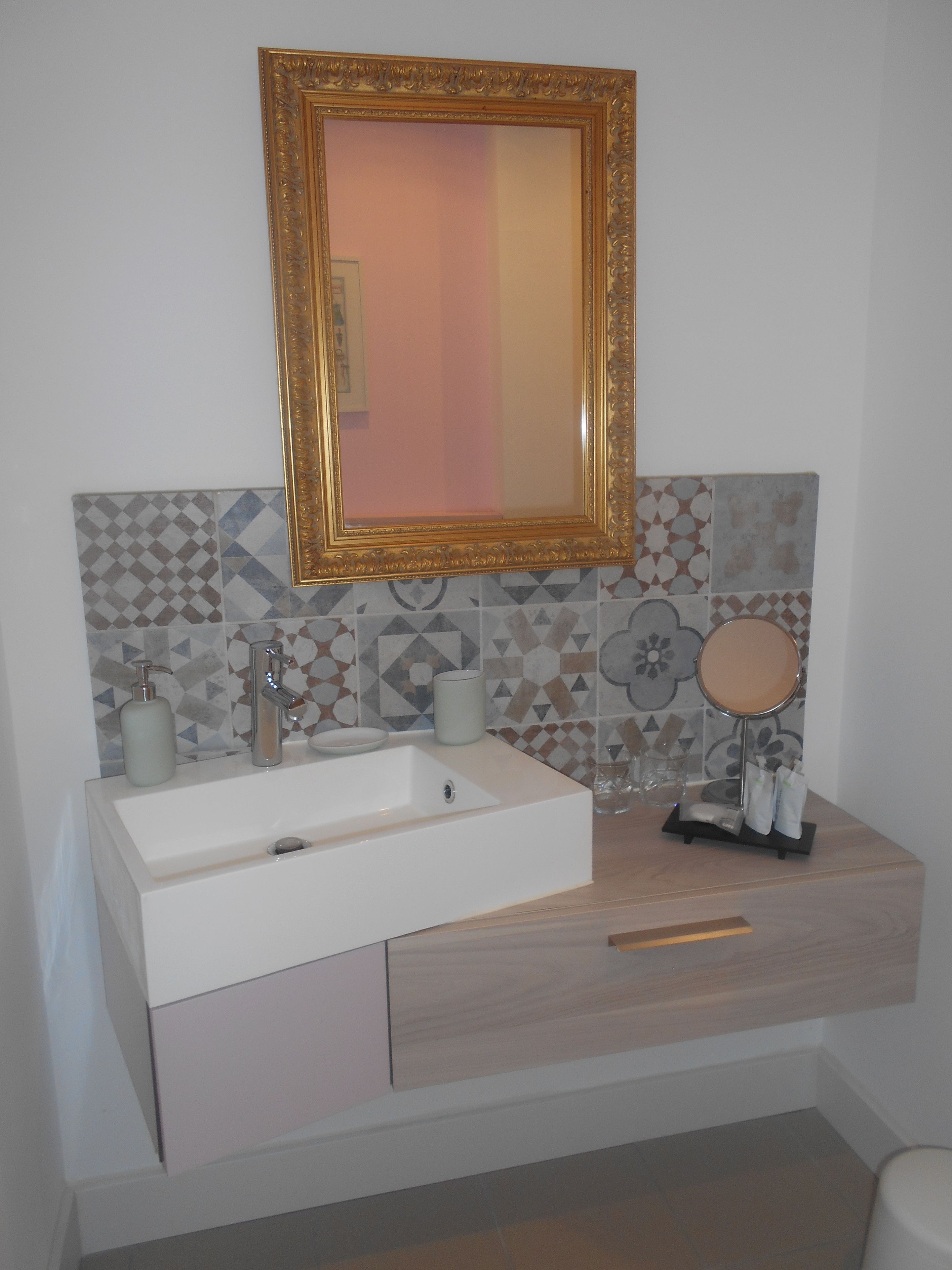 Ein Badezimmer mit einem weißen Waschbecken, einem spiegel mit goldener Rahmung und einer gefliesten Rückwand.
