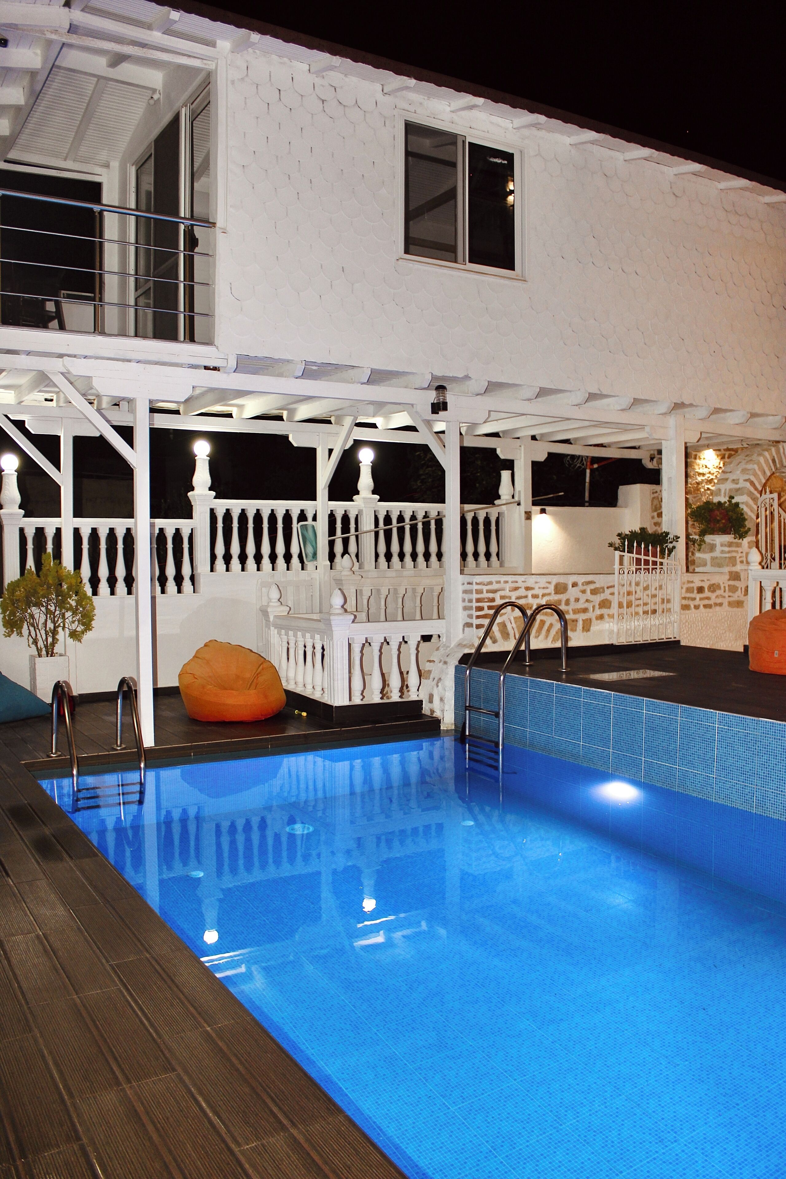 Une piscine avec une terrasse en bois, un bâtiment blanc doté d’un balcon et une clôture blanche.
