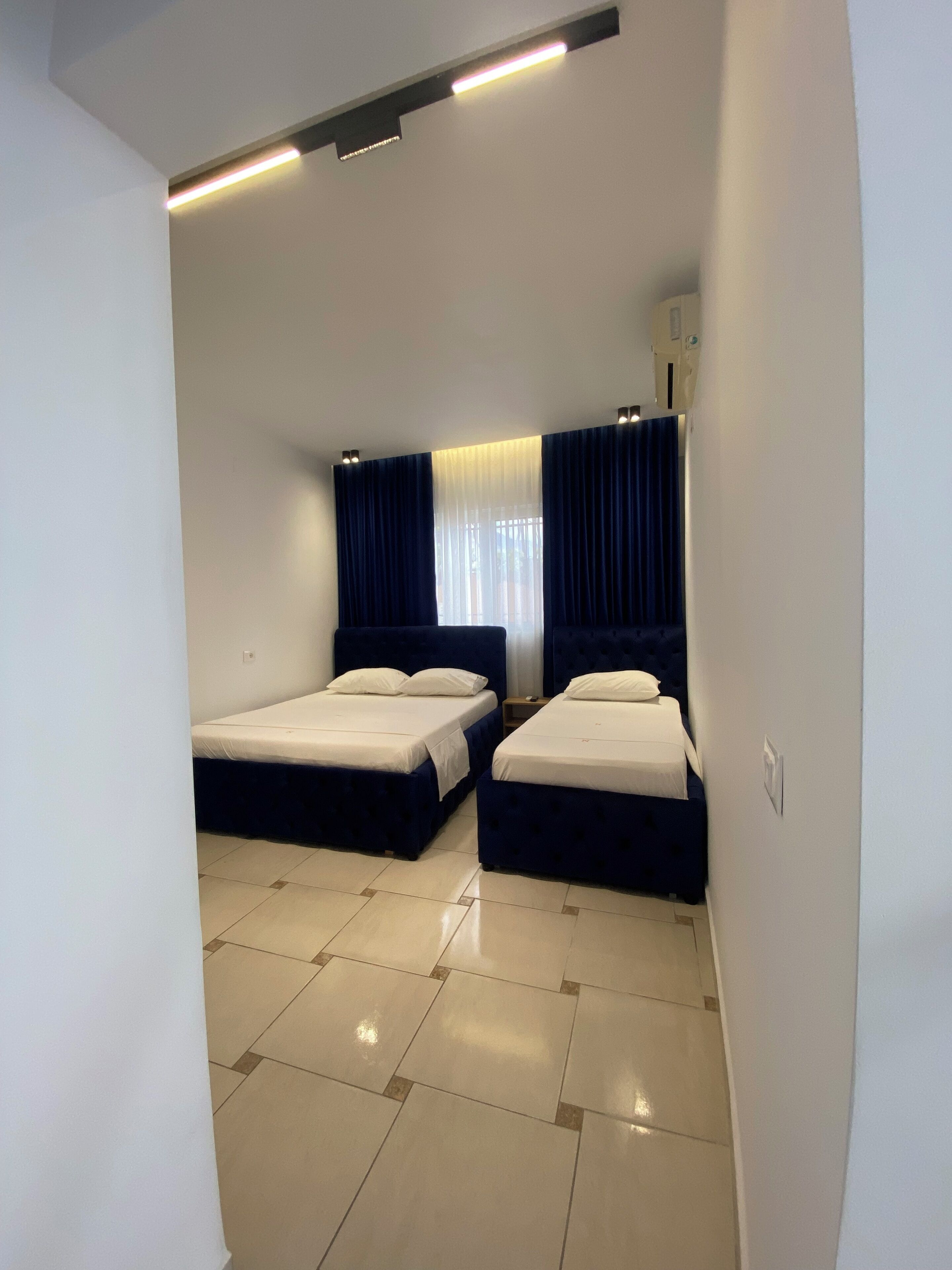 Habitación de hotel con dos camas, un ventanal con cortinas, una lámpara en el techo y un radiador.