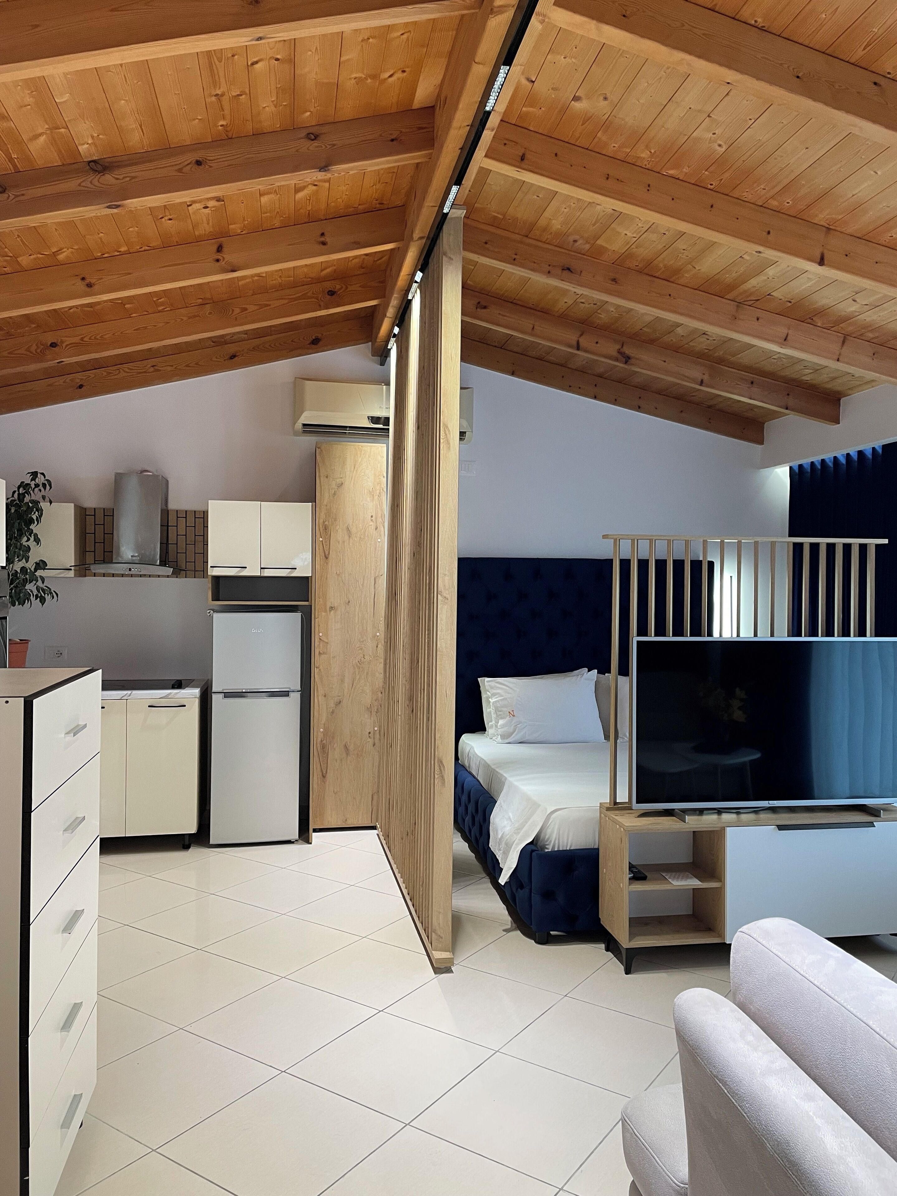 A compact room with a kitchenette, a bed with a TV, and a wooden ceiling.
