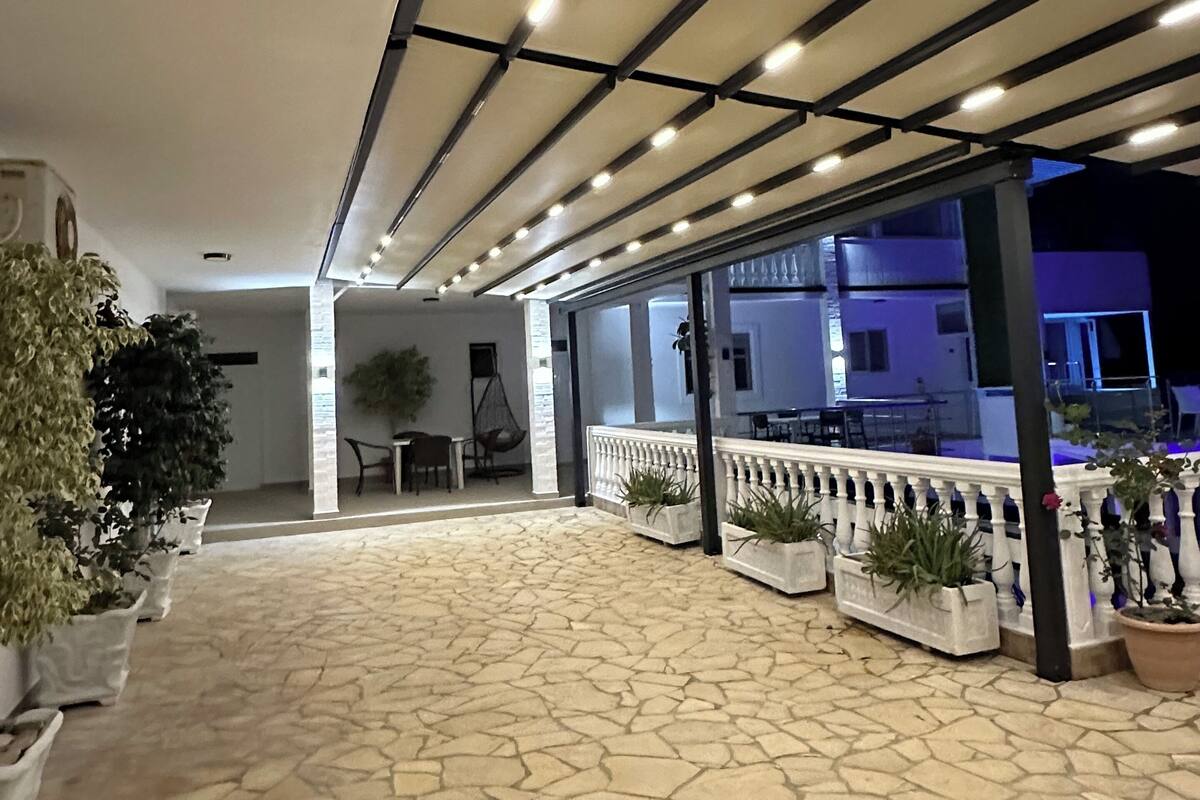 Uma área externa coberta com piso de pedra, plantas em vasos e um espaço de estar com mesas e cadeiras.