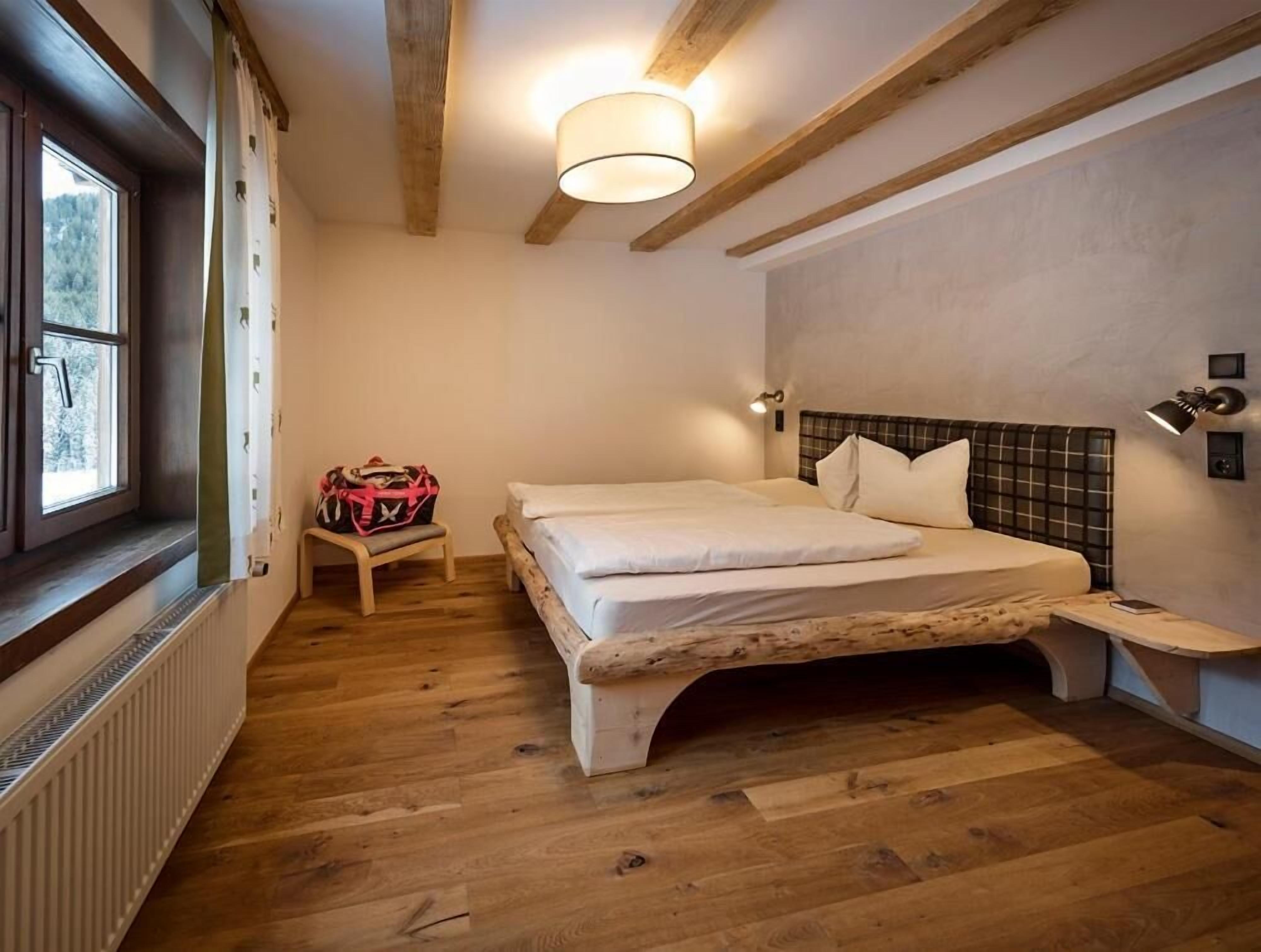 A bedroom with a wooden floor, a bed with a headboard, a bedside table, a lamp, a window, and a radiator.