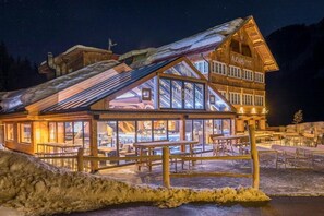 Una cabaña de madera con un cerramiento de cristal, mobiliario exterior y montañas al fondo.