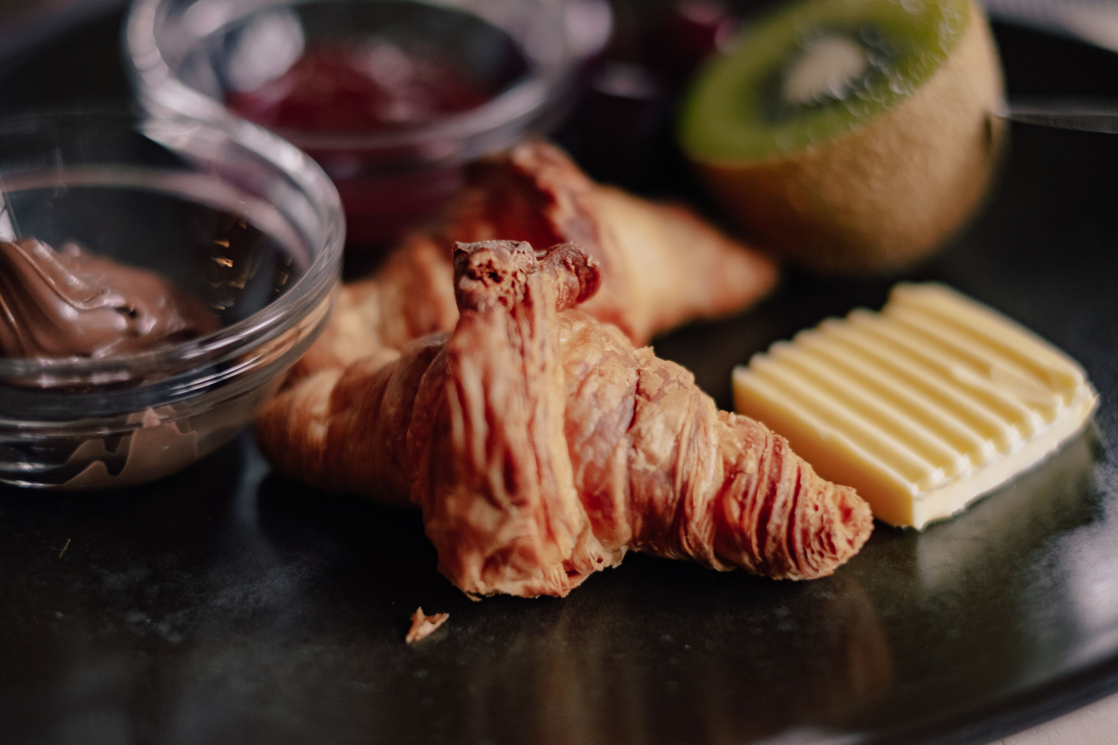 Un plato con un croissant, un kiwi, una porción de mantequilla y un cuenco pequeño de chocolate.