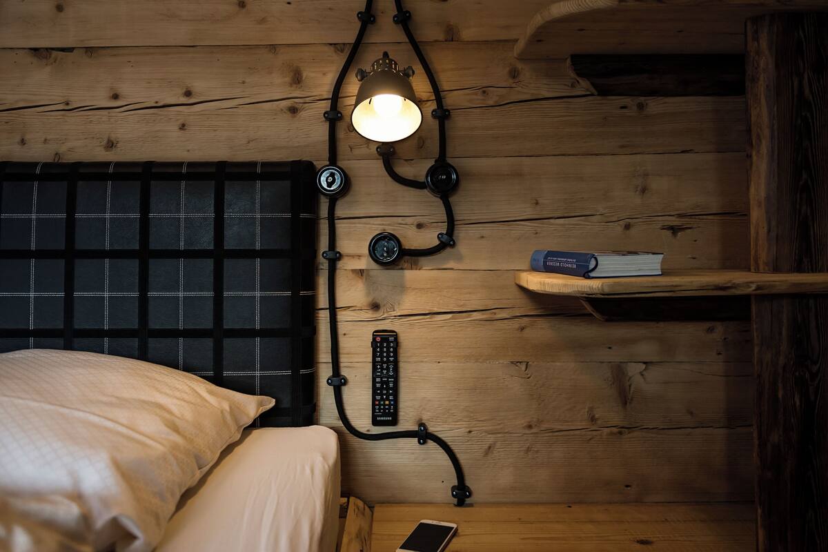 Um quarto de madeira com uma cama, uma mesa de cabeceira com um controle remoto, uma luminária e um livro em uma estante.
