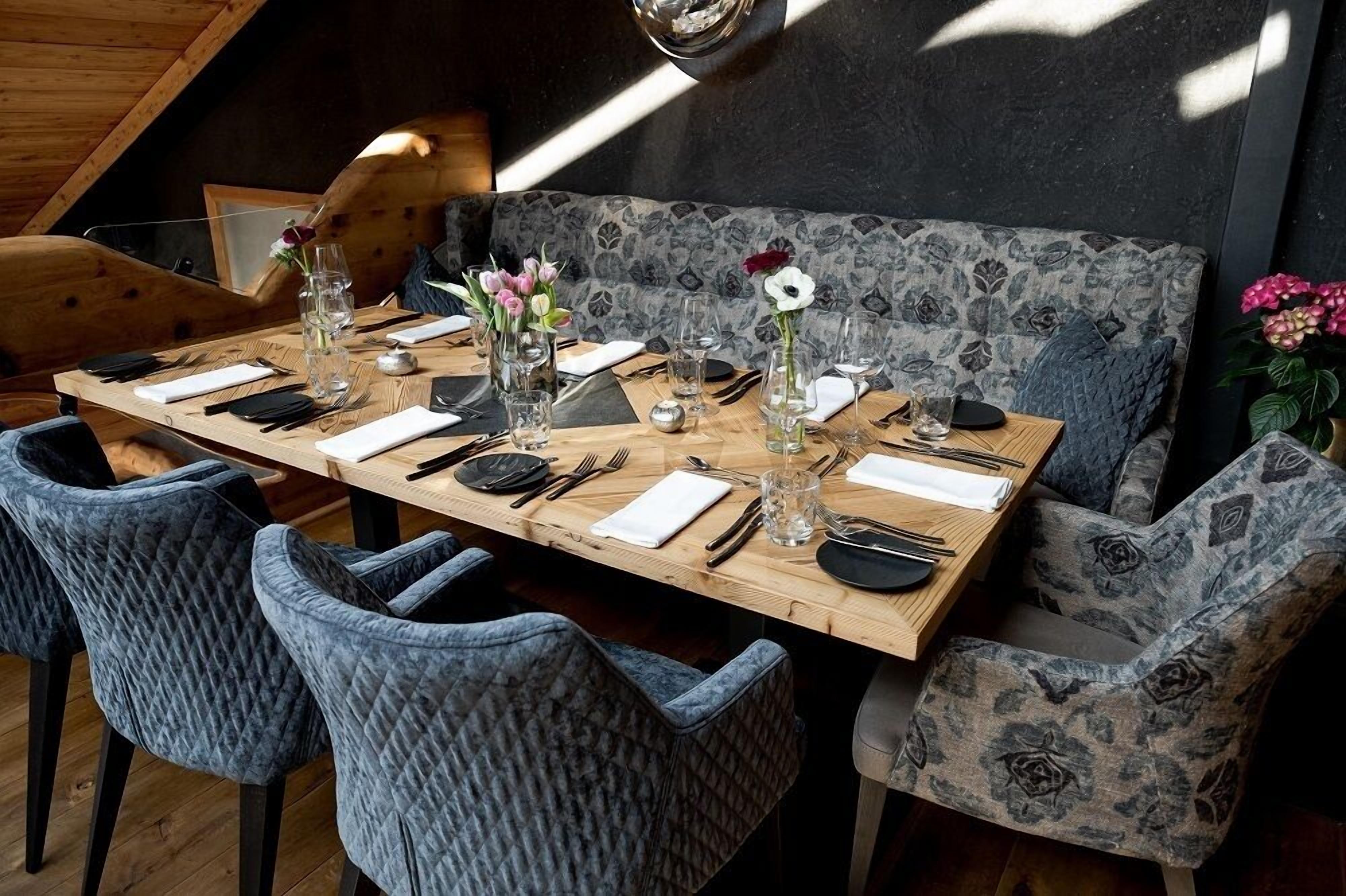 A dining area with a wooden table set for four, grey upholstered chairs, and a patterned bench in the background.