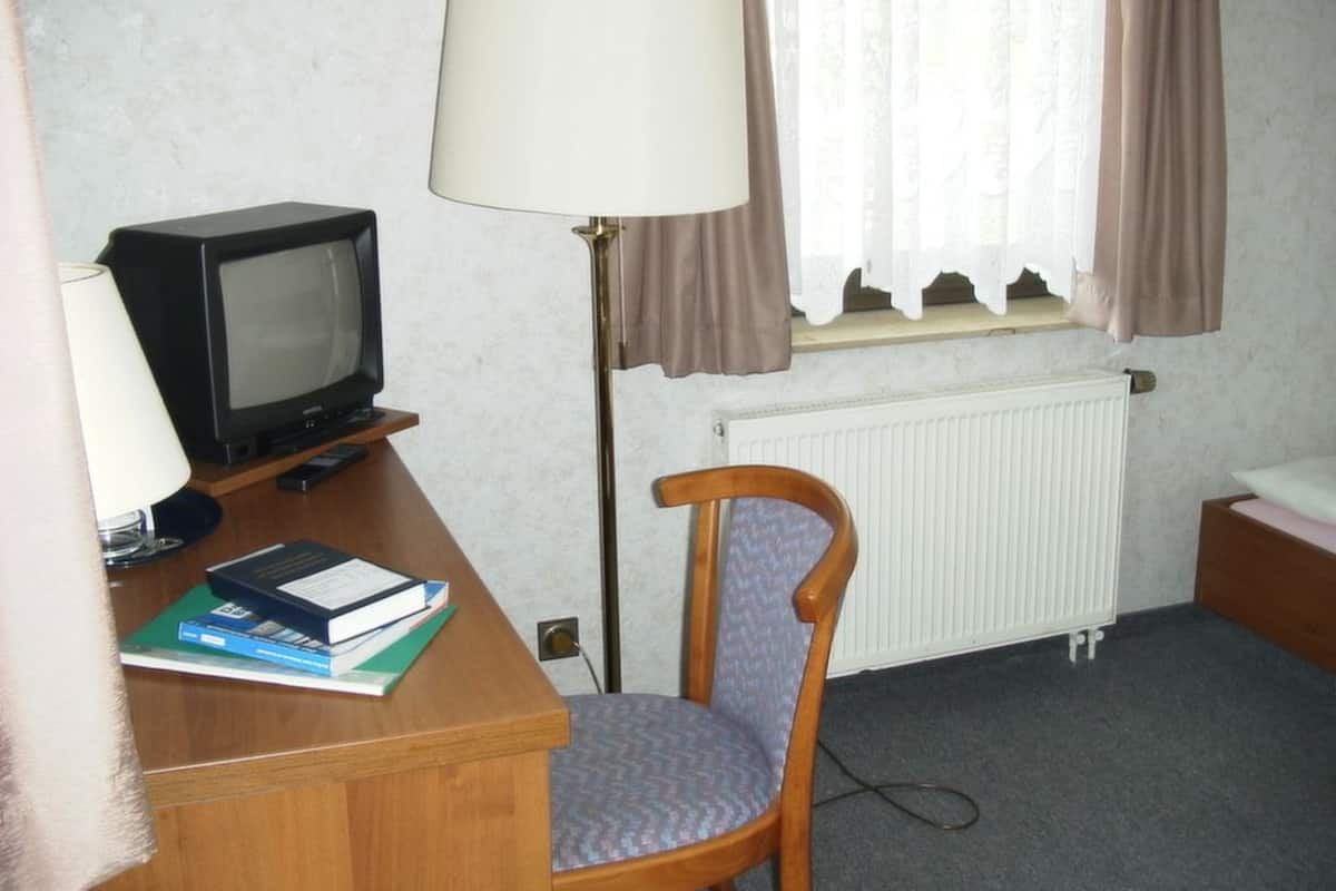 A hotel room with a wooden desk, a television, a chair, a radiator, and a window with curtains.