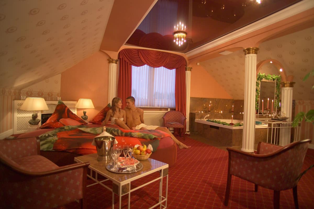 A hotel room with a bed, two chairs, a table with a fruit bowl, and a small table with a tray.