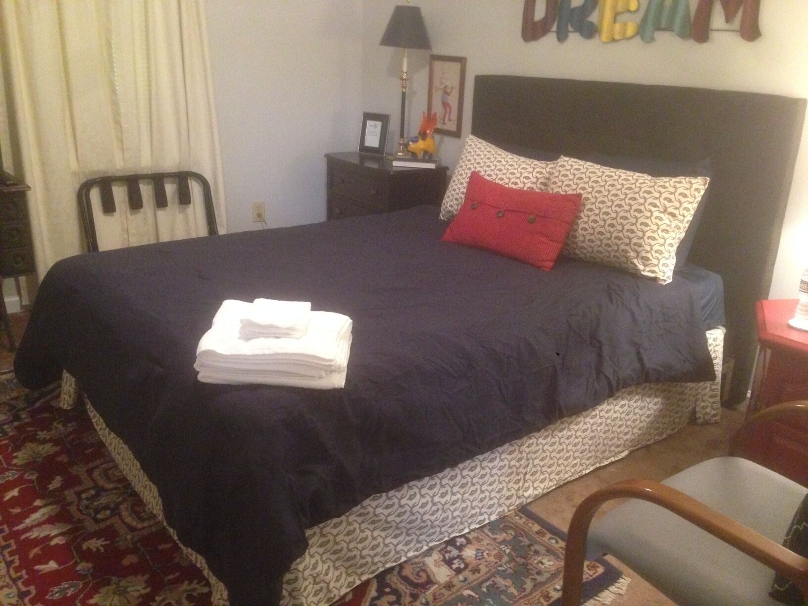 A neatly made bed with a dark blue bedspread and a red pillow, a small bedside table with a lamp, and a chair in a room with patterned carpet.