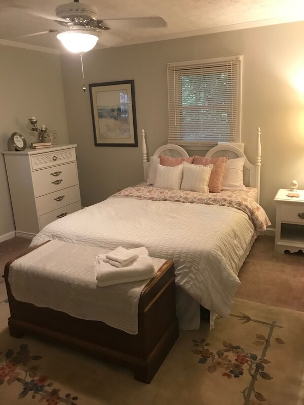 Une chambre à coucher comprenant un lit, une table de chevet, une commode, un ventilateur de plafond et une fenêtre avec des stores.
