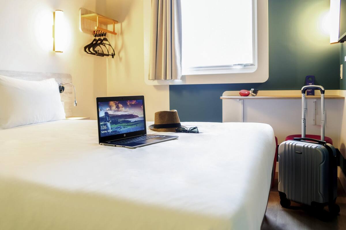 Habitación de hotel con cama, laptop sobre la cama, maleta, sombrero y una lámpara de pared.