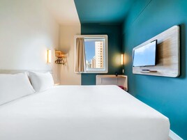 Una habitación de hotel con una cama, un televisor montado en la pared, un ventanal con vistas a edificios y una mesita con una lámpara.