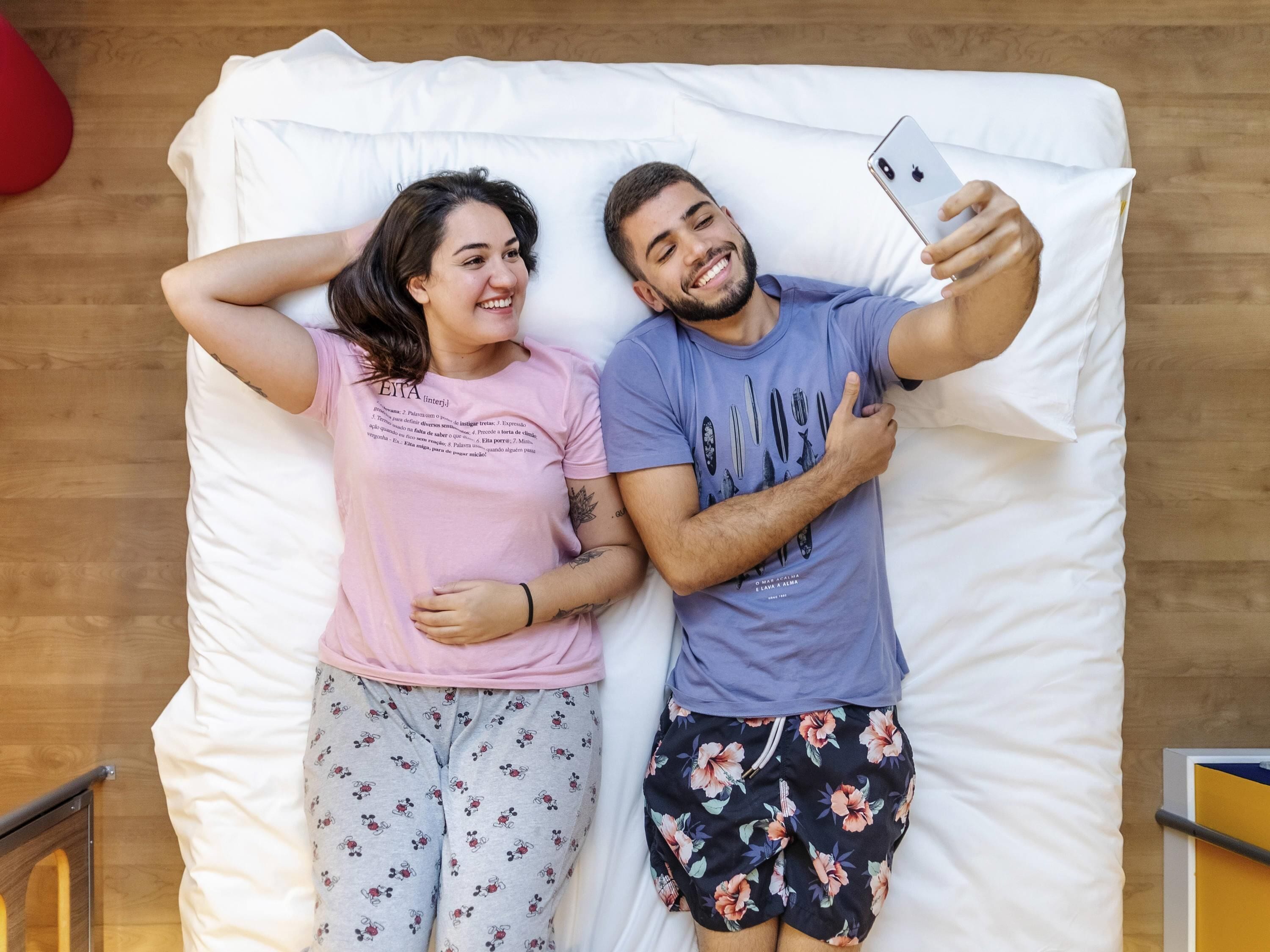 Un couple est allongé sur un lit, un smartphone est à proximité, le sol est recouvert de parquet et un objet rouge se trouve à l’arrière-plan.