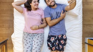 Ein Paar liegt auf einem Bett, ein Smartphone in der Hand, Holzboden und ein rotes Objekt im Hintergrund.