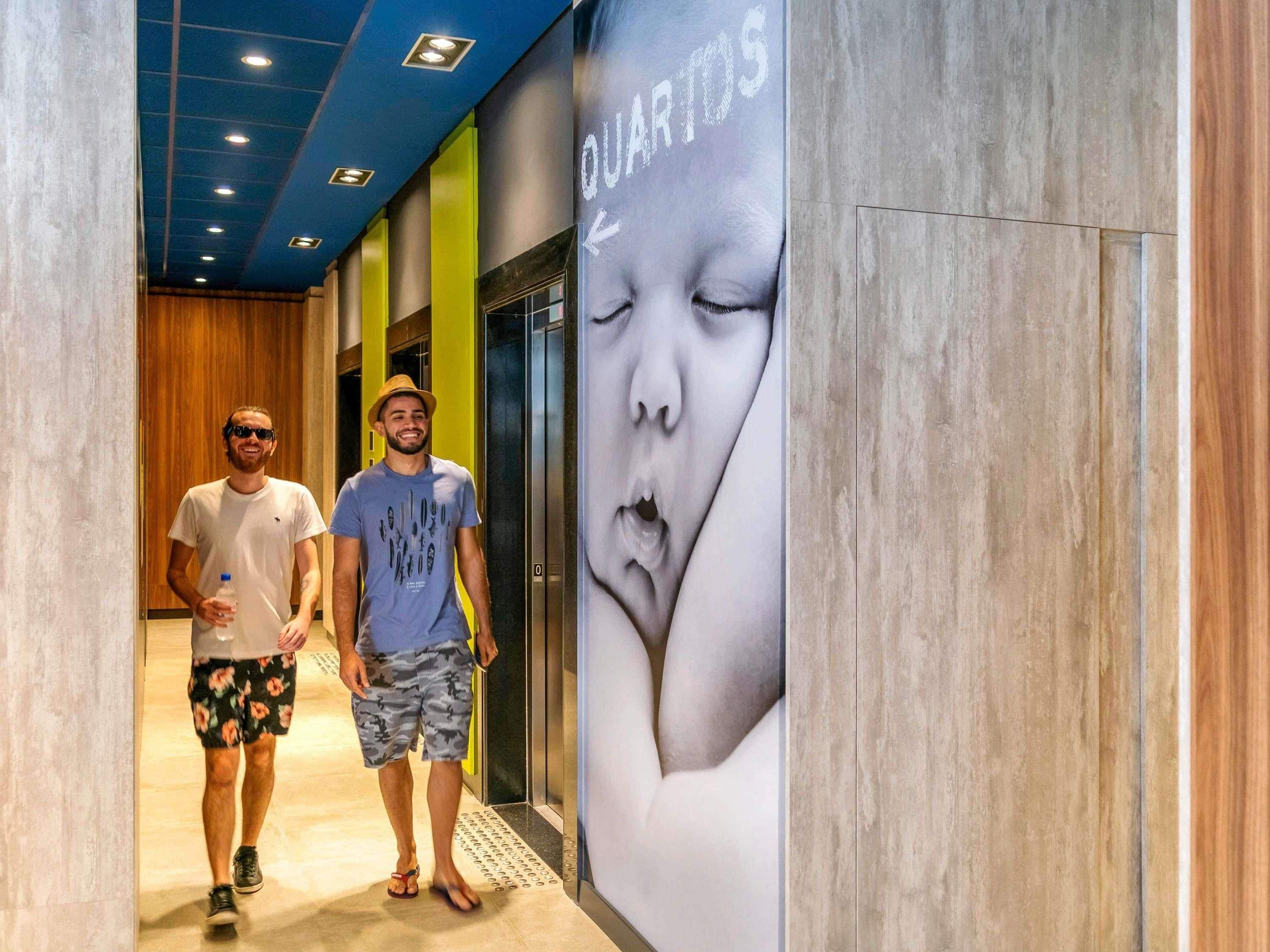 Two individuals walking in a hallway with a large advertisement featuring a baby's face and the word 'QUARTOS'.