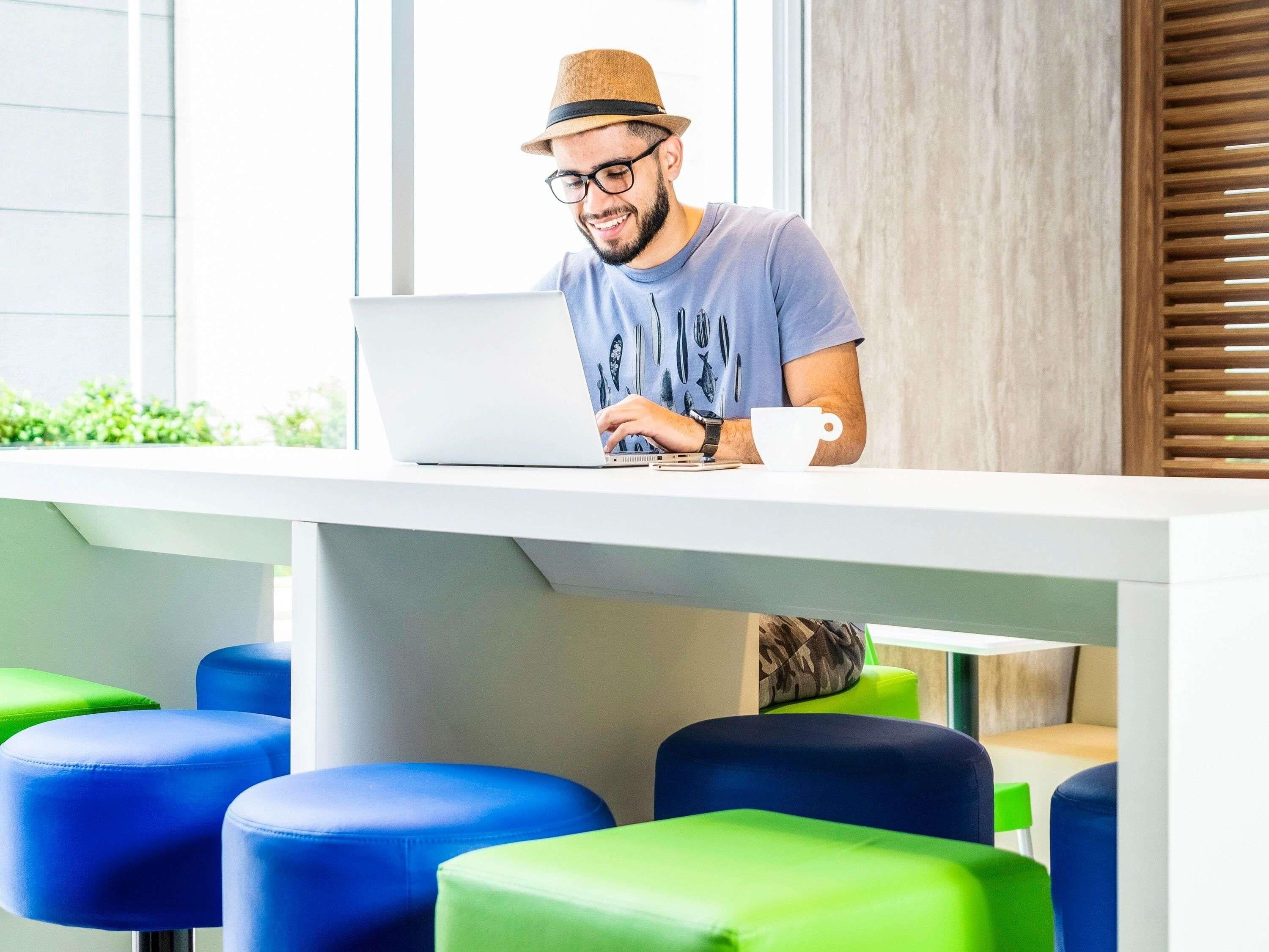 Una persona trabaja en una computadora portátil sobre una mesa blanca, con una taza a su lado, en una habitación con sillas azules y verdes.