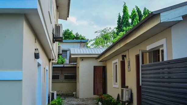 A narrow alleyway with a building on one side and a closed gate on the other. There are potted plants along the alley, and an air conditioning unit is visible on the wall. - The Emperor Place Annex (Lagos)