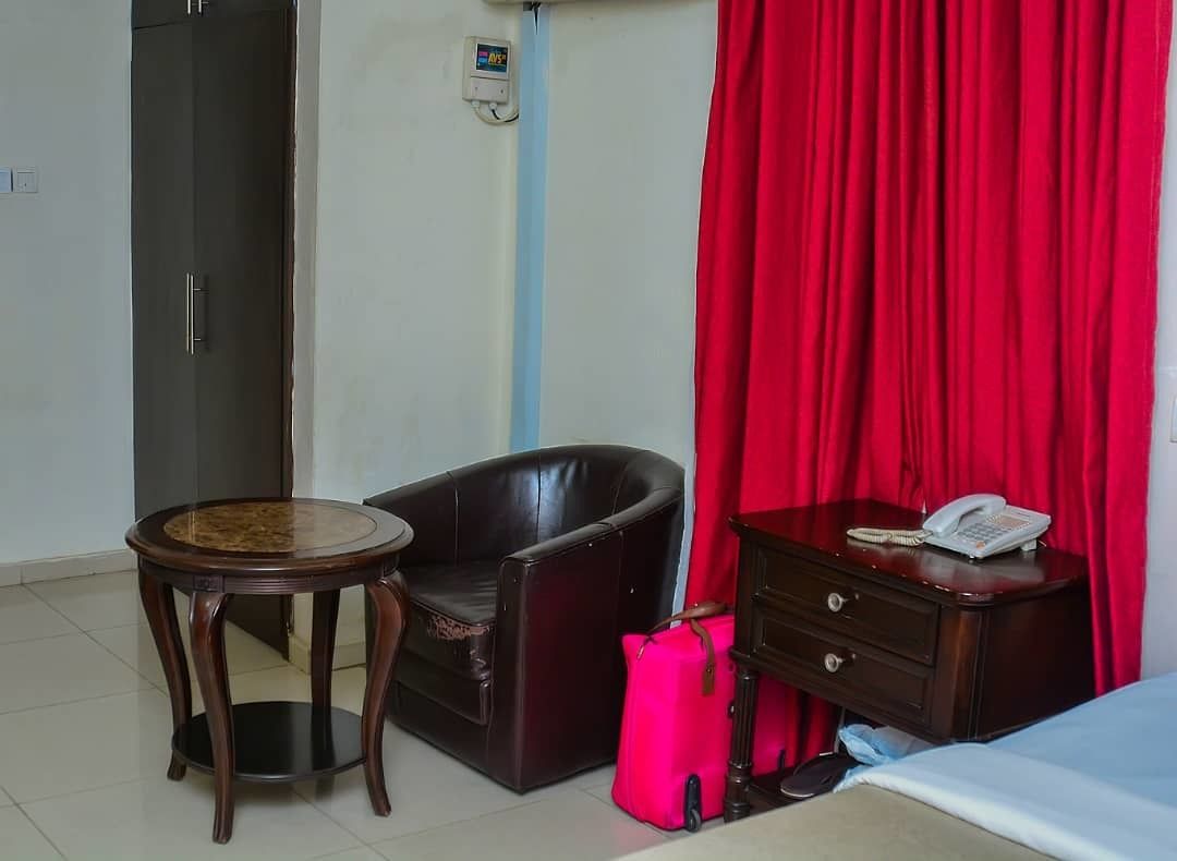 Una habitación de hotel con un sillón de cuero oscuro, una mesita de madera, un aparador de madera con un teléfono y una cortina roja brillante.