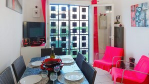 Zona pranzo con tavolo apparecchiato per quattro persone, televisione a parete e vista attraverso una finestra con persiane.