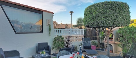 An outdoor dining area with a table set for four, surrounded by wicker chairs. There is a white wall with a window and a small potted plant. A large tree and a white fence are visible in the background.