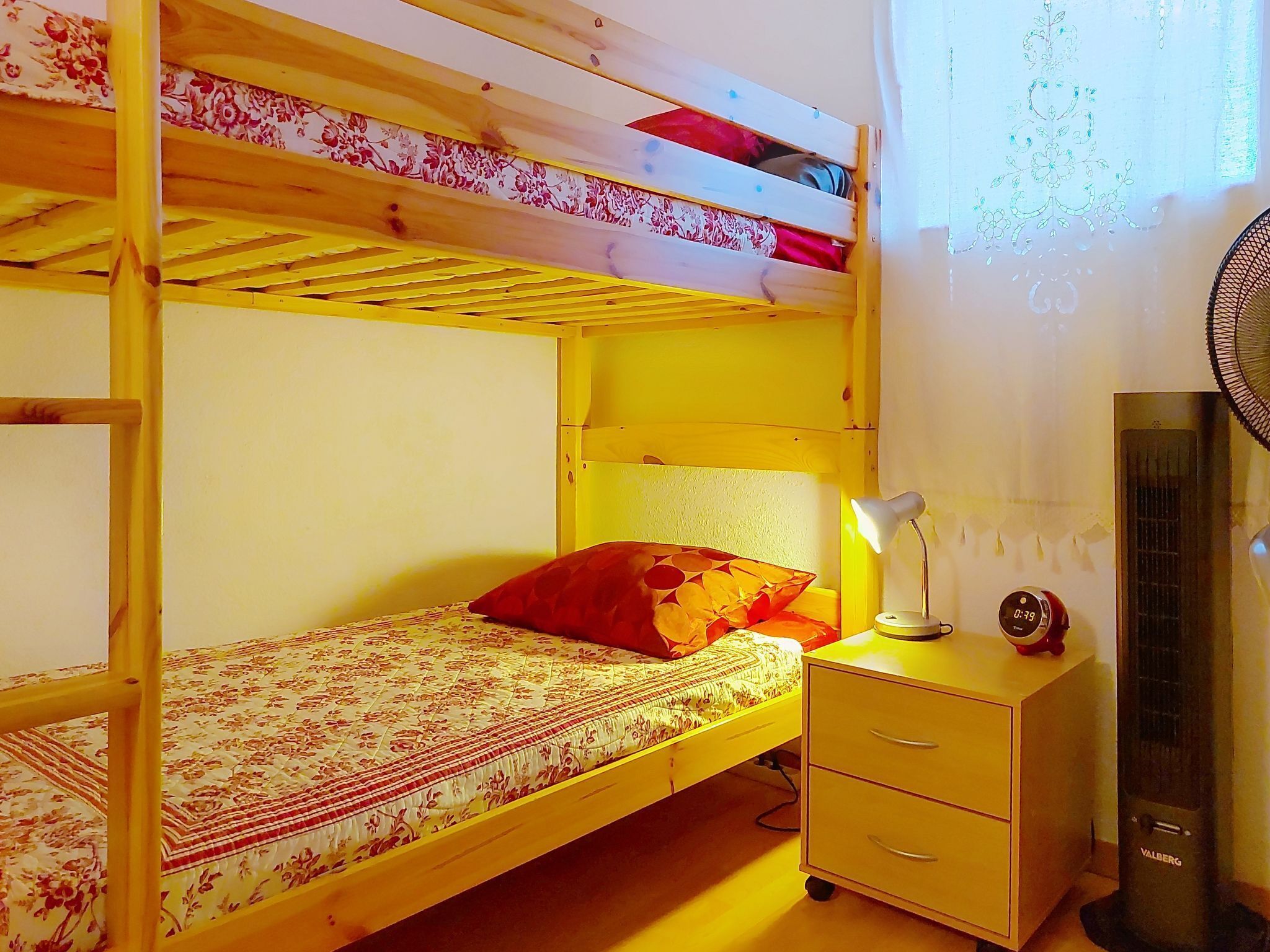A bunk bed with a floral-patterned bedspread, a red pillow, a bedside lamp, and a small white nightstand with drawers.