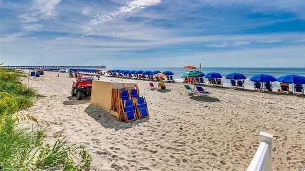 Una playa con hileras de sombrillas azules y sillas, una torre de salvavidas y un muelle al fondo.