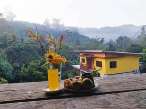 A yellow house with a balcony, a wooden table with a glass of yellow liquid and a plate of fruits, and a view of a lush green landscape. - Bondoi Lampang (Mae Tha)